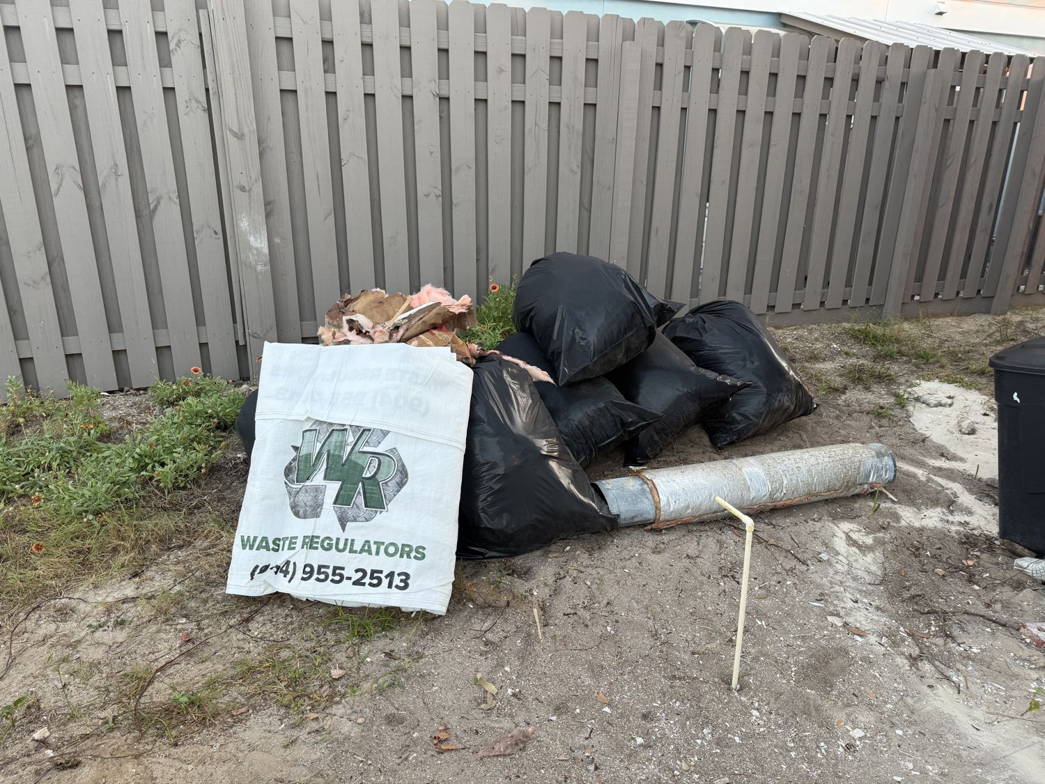 A filled Waste Regulators dumpster bag beside a fence, ready for pickup